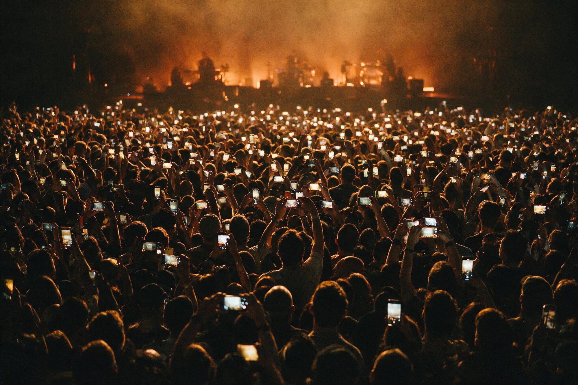 A concert crowd from behind, hundreds of raised phones recording the stage